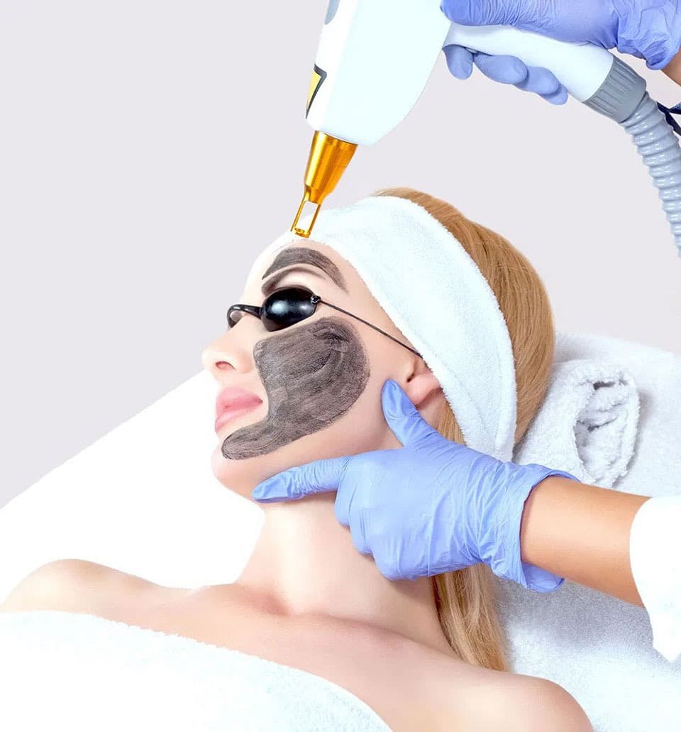 aesthetician placing hollywood facial device on forehead of woman with carbon face mask wearing eye covers